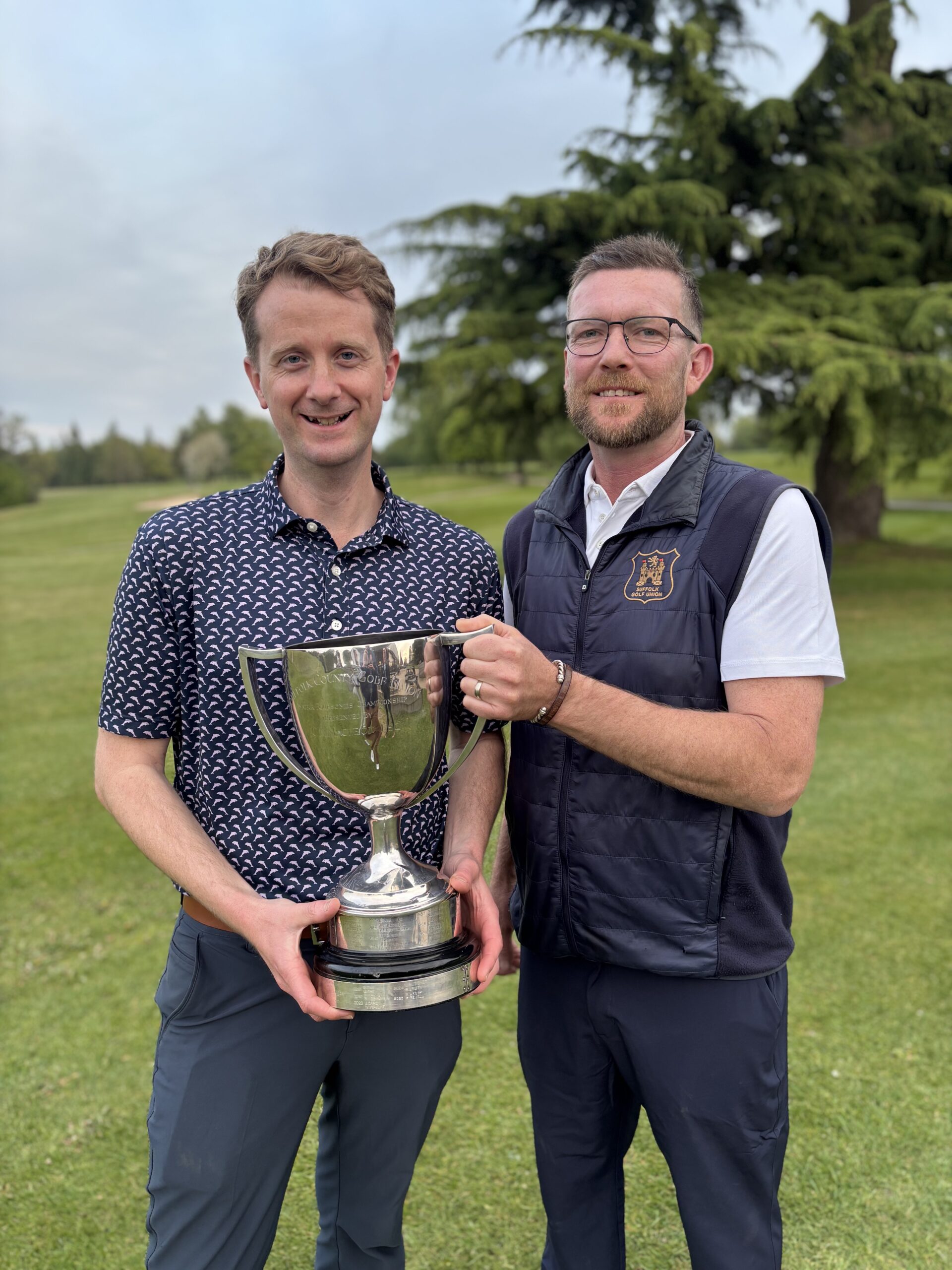 A photo showing 2026 Suffolk Amateur Foursomes Champions Daniel Hedges and Russel Oakey holding the winning cup on a golf course.